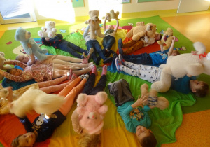 Grupa dzieci leży na chuście animacyjnej z skierowanymi nogami do środka, podrzucają misie.